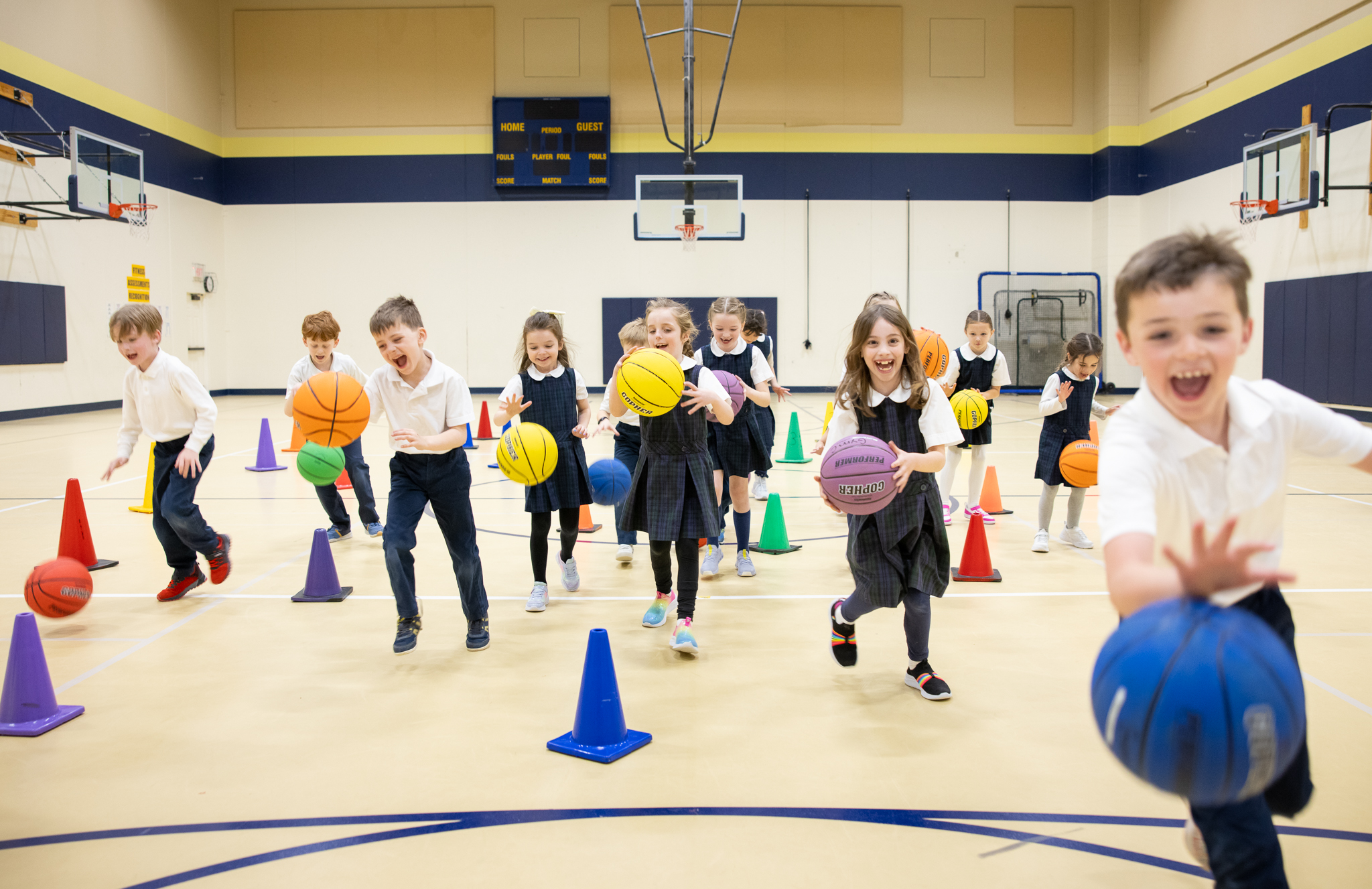 Lower School students dribbling basketballs across the gym, weaving through cones and laughing.