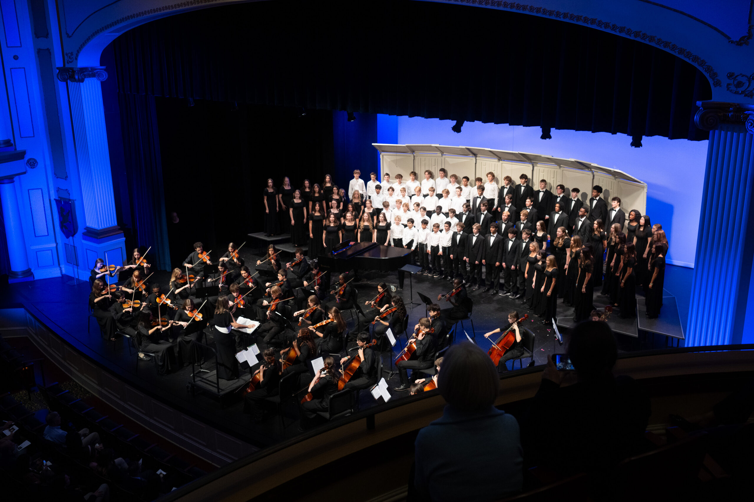 Providence Academy choir and string orchestra performing together on stage in formal black concert dress under blue stage lighting